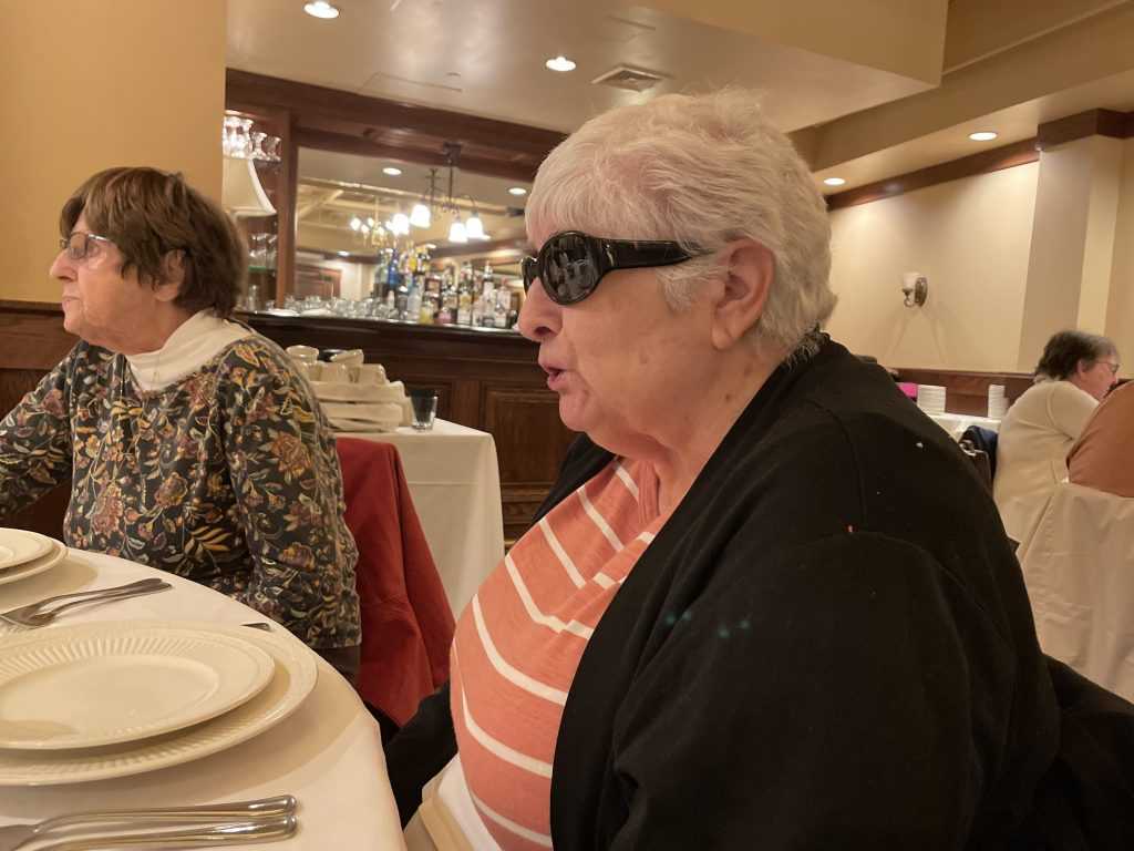 Two women, one wearing sunglasses and one wearing eyeglasses, sit a table talking to people across from them.