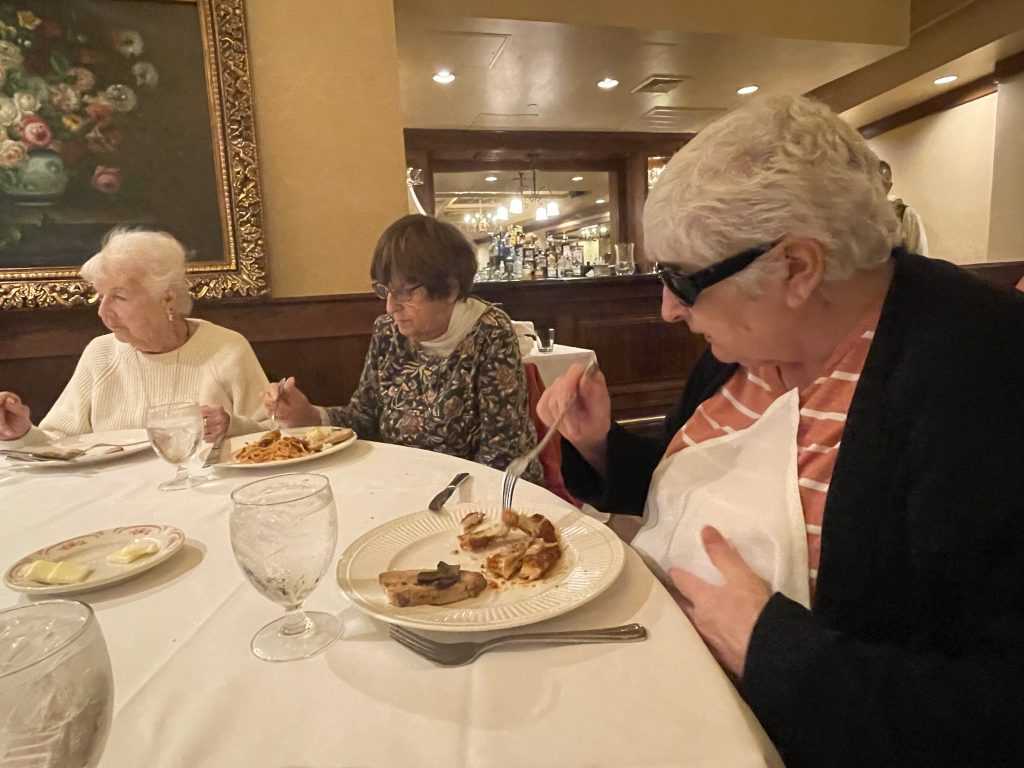 Three women sit at a table holding forks and eating food off their plates.