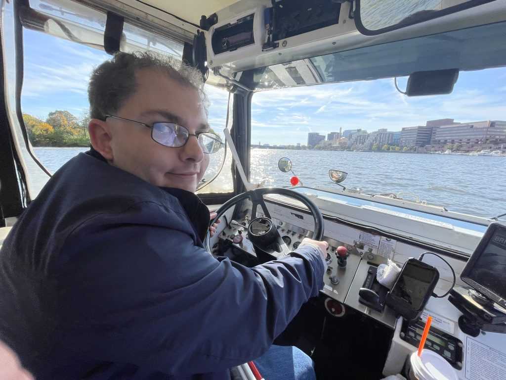 A male sits behind the steering wheel of a duck boat as it drives in the water.