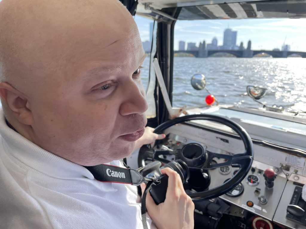 A male sits behind the steering wheel of a duck boat as it drives in the water. Tall buildings can be seen in the background.