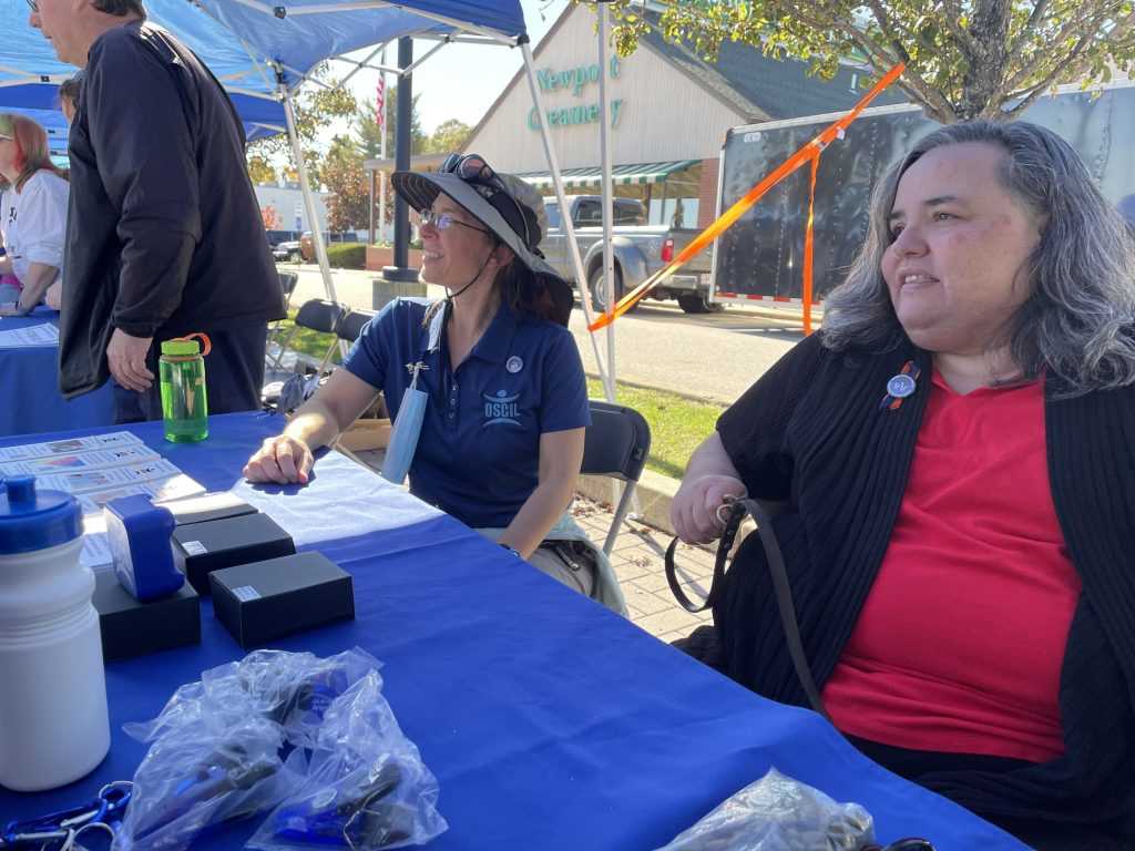 Two staff members from Ocean State Center for Independent Living (OSCIL) sit behind a table offering information to White Cane day participants.