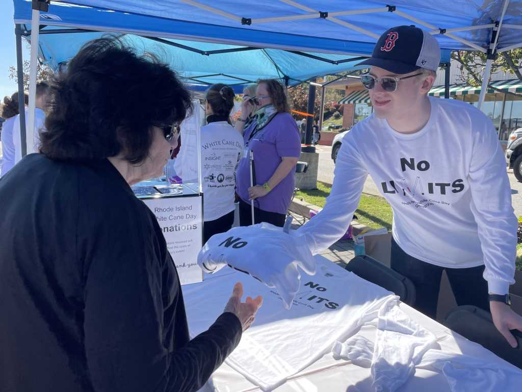 Rhode Island Parents of Blind and Visually Impaired Children (RIPBVIC) volunteer Jackson, wearing a baseball hat and a white t-shirt with the words "No Limits" printed on it, hands a folded t-shirt to a woman on the opposite side of the table.