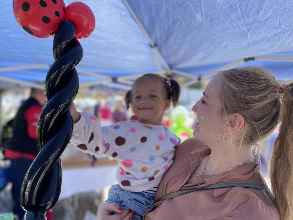 A little girl in her mom's arms holds black and red balloons that have been twisted into the shape of a ladybug.