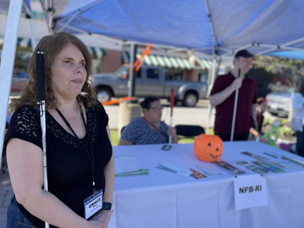 Grace, president of the National Federation of the Blind of Rhode Island, stands in front of the NFB informational table. Two other chapter members are behind the table.
