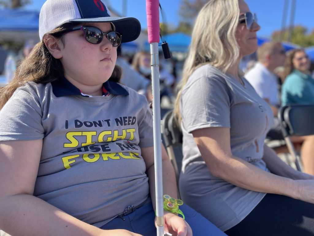 A young girl wearing a baseball cap, sunglasses, and a t-shirt that reads, "I don't need sight to use the force," sits in a chair holding her white cane with a pink grip in front of her.