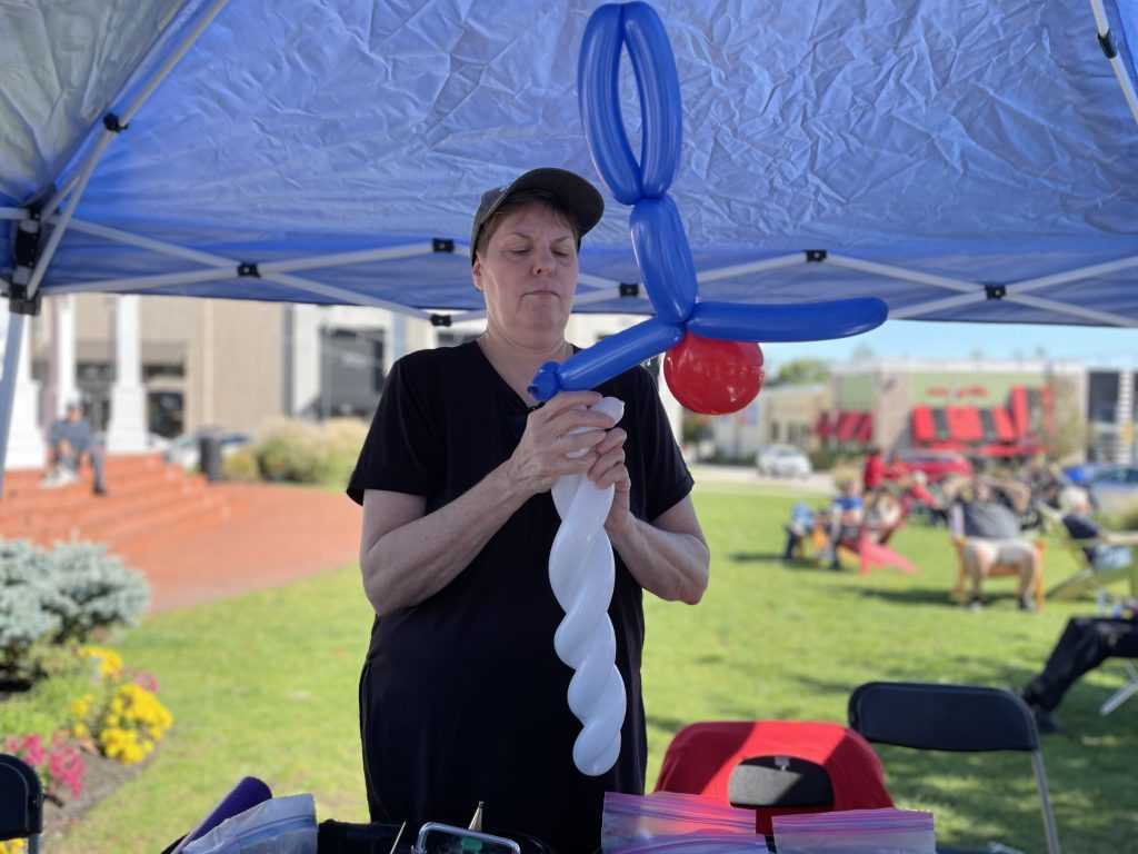 A balloon artist wearing a black shirt and baseball hat, twists blue and white balloons together.