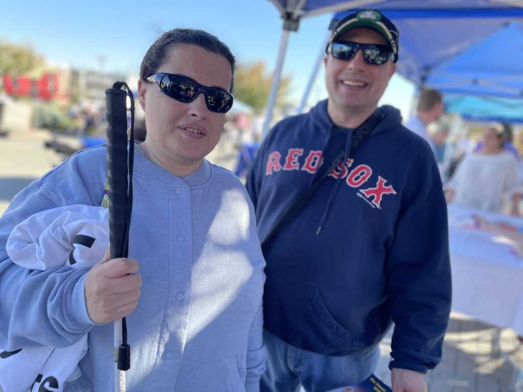 White Cane Day participant Angie, wearing sunglasses and holding a cane in one hand, looks towards the camera. Her husband, Peter, wearing a Red Sox sweatshirt, stands behind her.