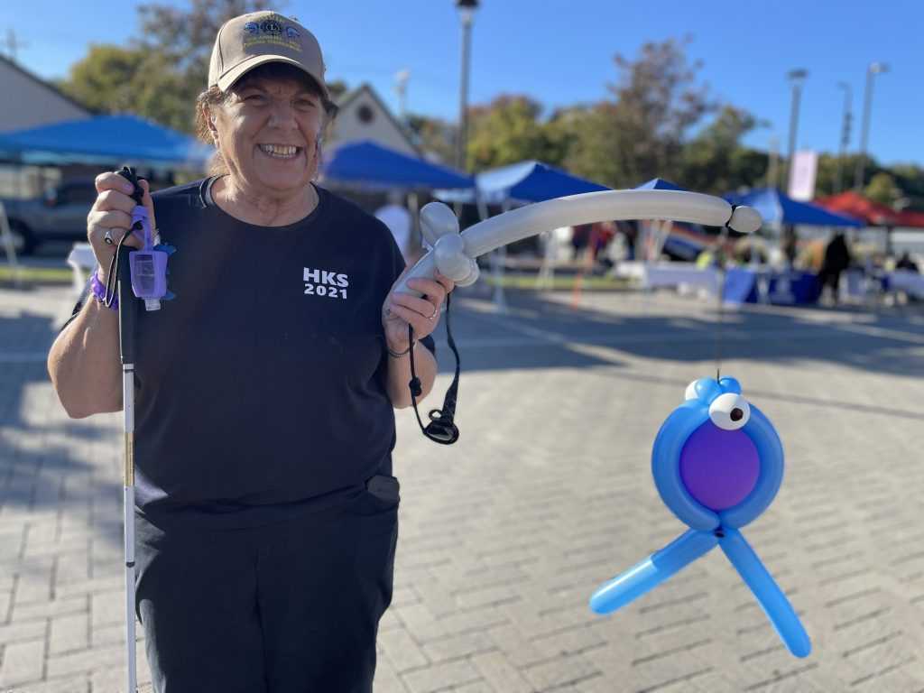 White Cane Day event participant Janice, wearing a baseball hat and a blue shirt, stands holding her cane in one hand and balloons that have been twisted into the shape of a fish hanging from a fishing pole in her other.