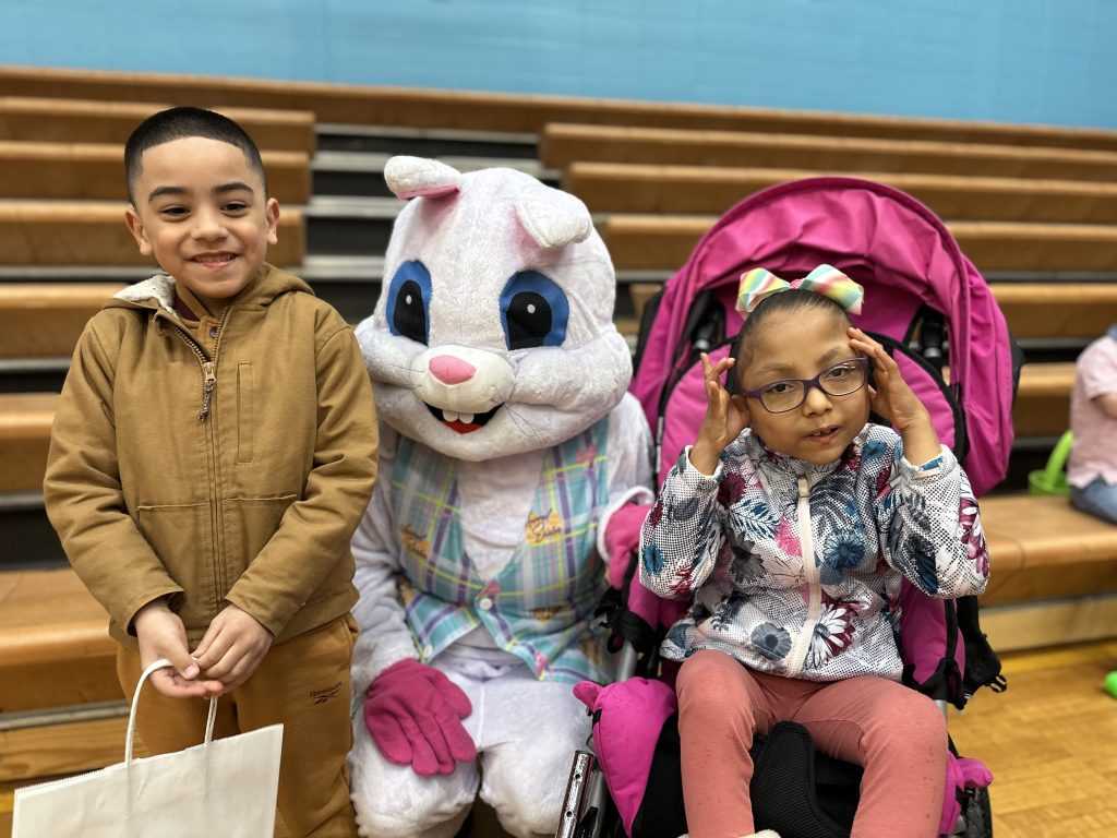Two kids, one in a stroller, sit on either side of the Easter Bunny.