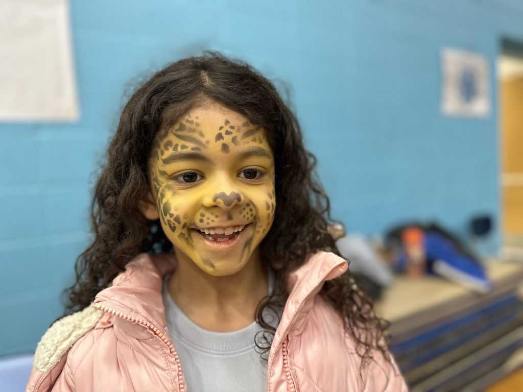 A girl with long brown hair with her face painted yellow with a cheetah spots, looks at the camera.