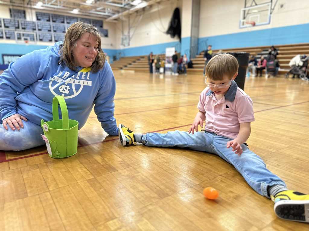 A boy sits on the floor with his legs spread open, watches, an orange plastic egg as it rolls towards him, while his mother looks on.