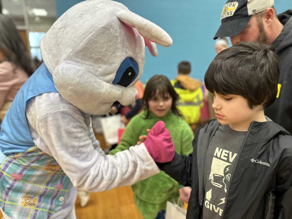 A young boy with dark hair and a black jacket shakes hands with the Easter Bunny.