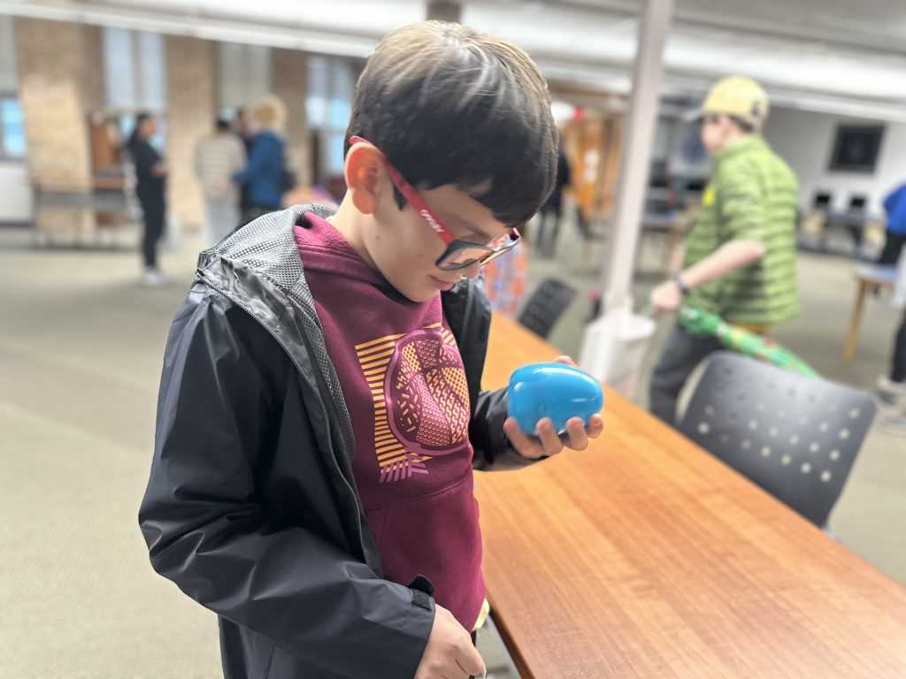 A boy wearing a red shirt with a black jacket and red and black frames glasses looks closely at a blue beeping egg.