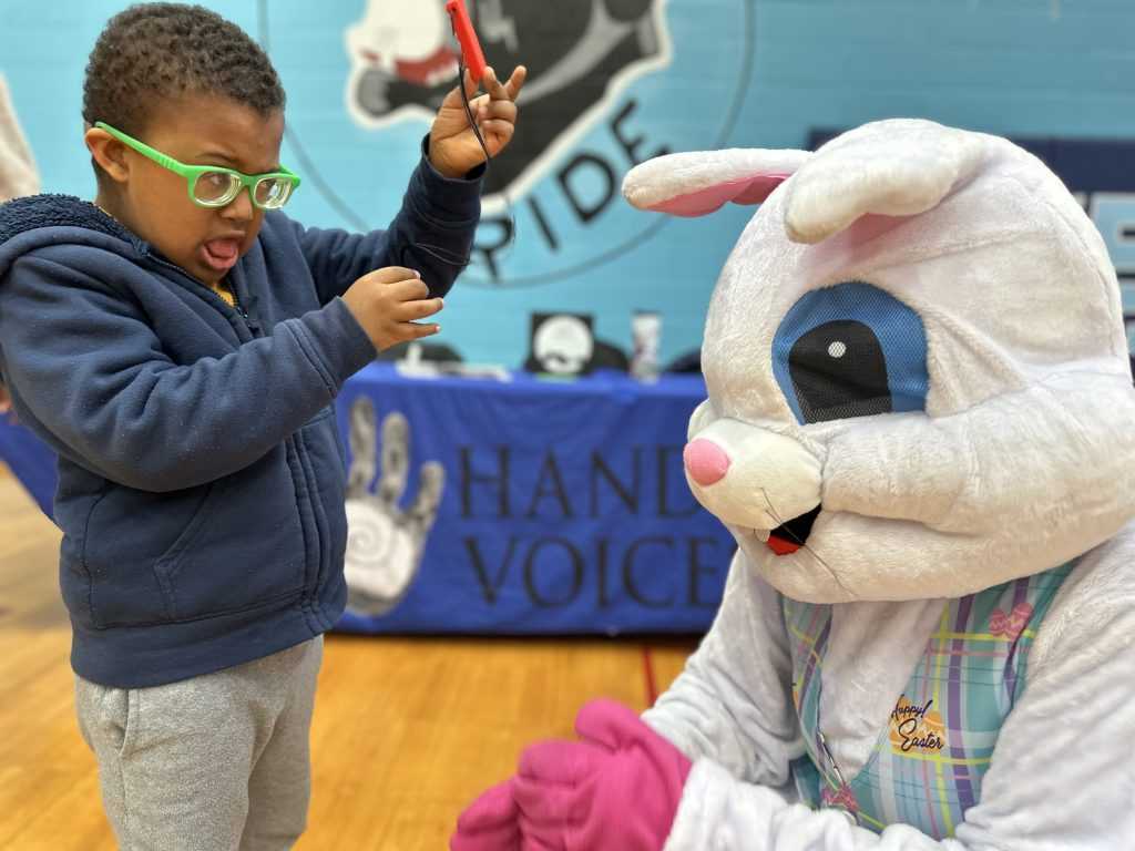 A boy wearing a blue hoodie and green frame glasses shows a toy to the Easter Bunny who is kneeling in front of him.