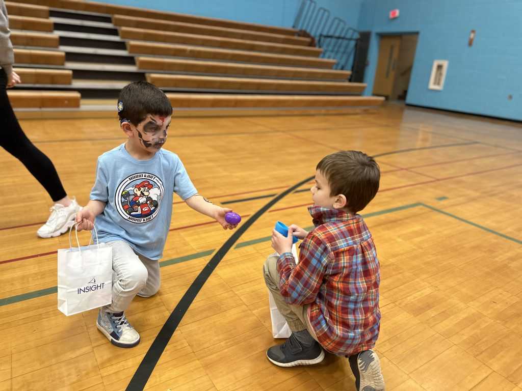A boy wearing a blue shirt with his face painted, hands a purple egg to another boy wearing a plaid shirt. Both are kneeling on a wood gym floor.